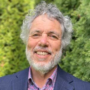 Smiling older man with curly gray hair and beard, wearing a blue blazer and patterned shirt, stands outside in front of green foliage—his warm demeanor reflecting the patience and wisdom that help him teach anyone to read.